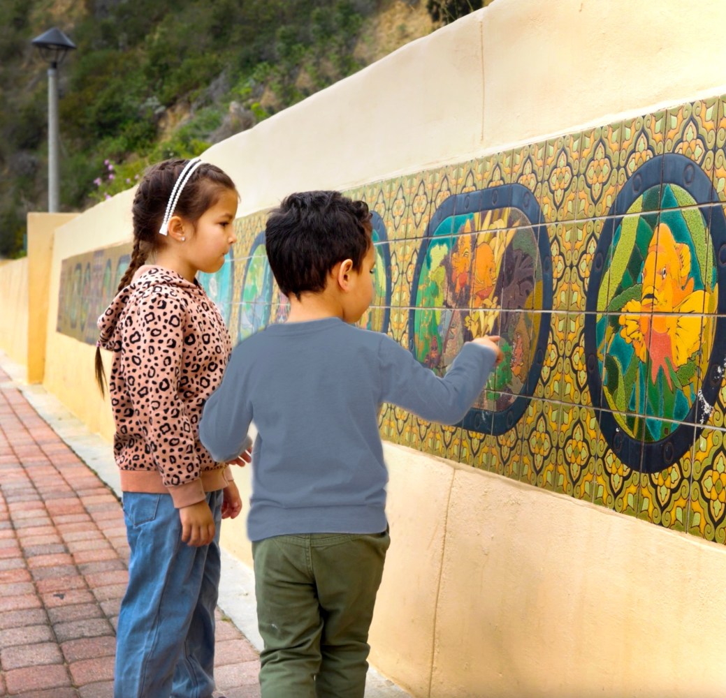 Two children observe colorful tile art on an outdoor wall.