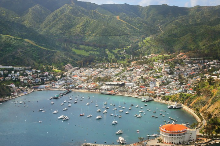 Coastal town with boats, hills, and a large round building by the water's edge.