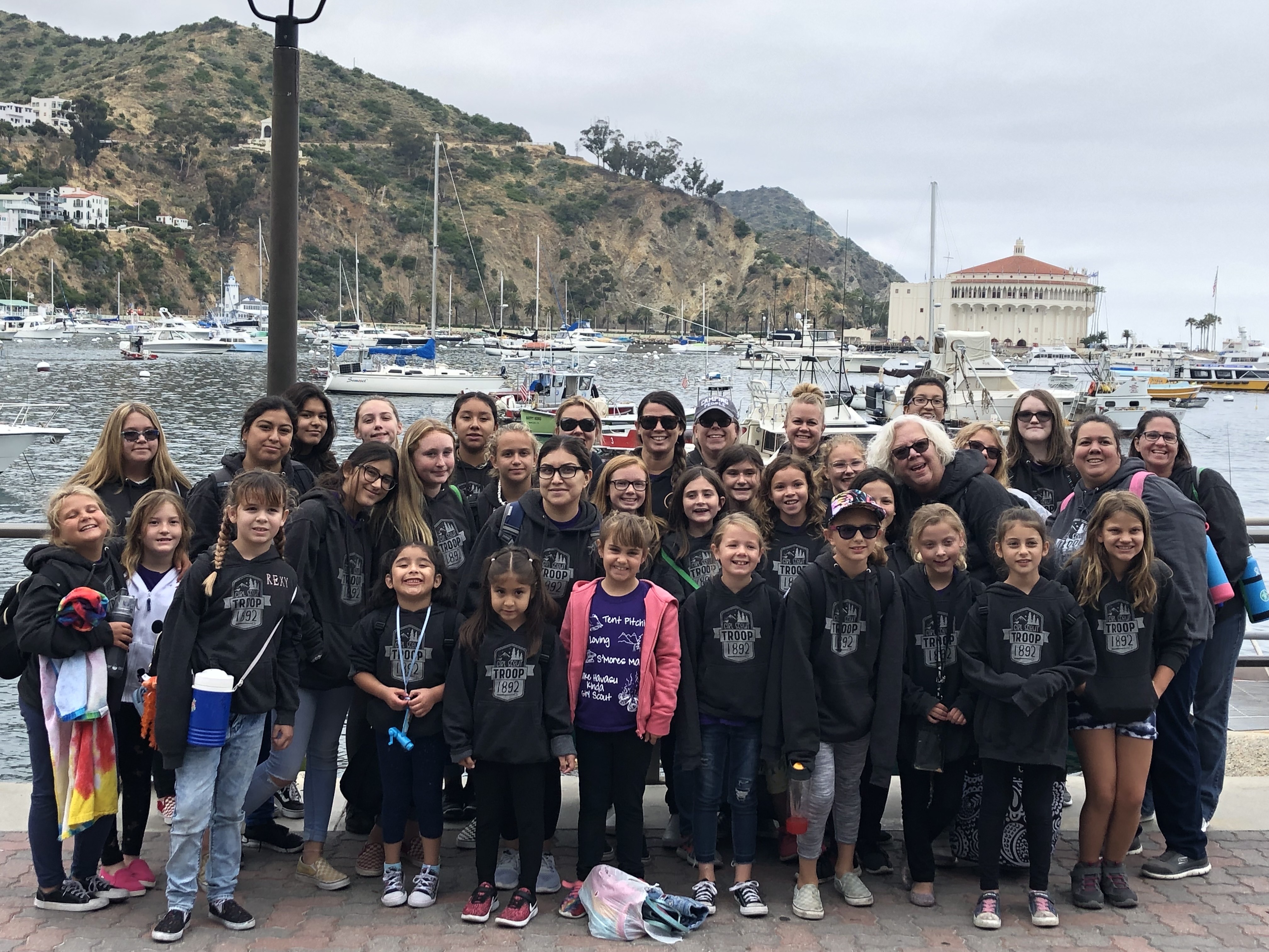 Group of Girl Scout Troops wearing matching hoodies posing by Avalon Bay with boats, hillside, and the Casino Building behind them.