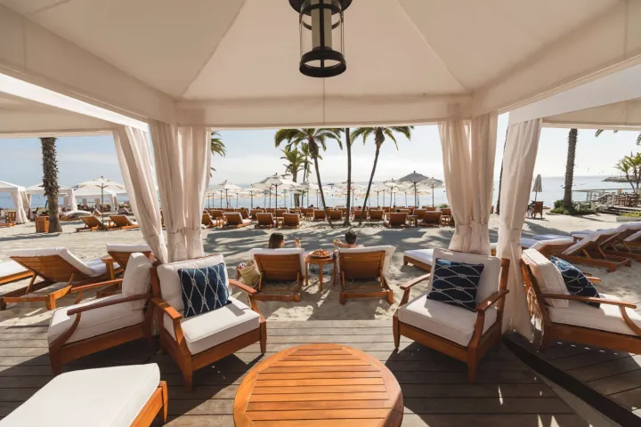 Beach cabana with loungers, table, and view of palm trees, umbrellas, and ocean.
