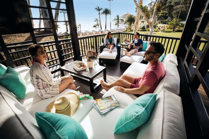 Four people relax in a gazebo with snacks, overlooking a scenic beach with palm trees.