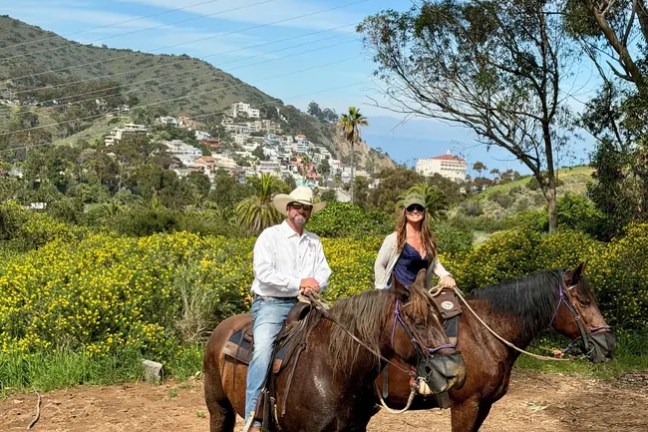 Two people on horseback in a scenic, hilly area with trees and buildings.